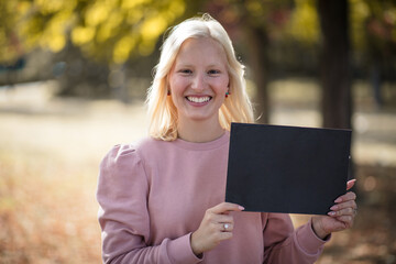 Smiling blond woman in the park holding black board. Empty space. Space for copy.