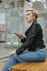 short haired caucasian woman wearing leather jacket and jeans sitting alone on  metro station with smartphone in hands waiting for train to come. Image with selective focus and noise effect