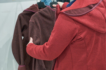 Caucasian woman chooses fleece burgundy jacket with hood jacket in store to buy. Concept of buying...