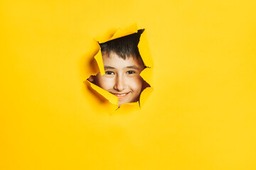 Closeup of the smiling face of a little Caucasian boy looking through a torn hole in a colored yellow paper wall.