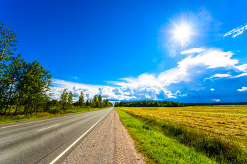 Fototapeta premium Straight country road passing through a field illuminated by the sun's rays. View from the side of the road