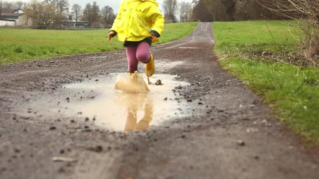 Close-up of little toddler girl wearing yellow rain boots and walking and running during sleet on rainy day. Cute child in colorful clothes jumping into puddle, splashing with water, outdoor activity