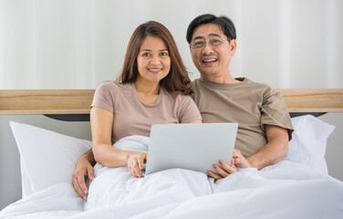 Obraz na płótnie Canvas happy middle age Asian couple dressed casually sitting relaxed on a bed at home enjoy looking at a laptop computer. Warmth, delightful family concept