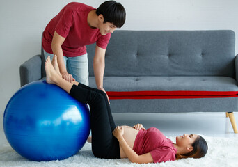 happy handsome young Asian man sitting looking at a pregnant woman happily. Active husband encourages his wife to do exercise with a pilates ball at home