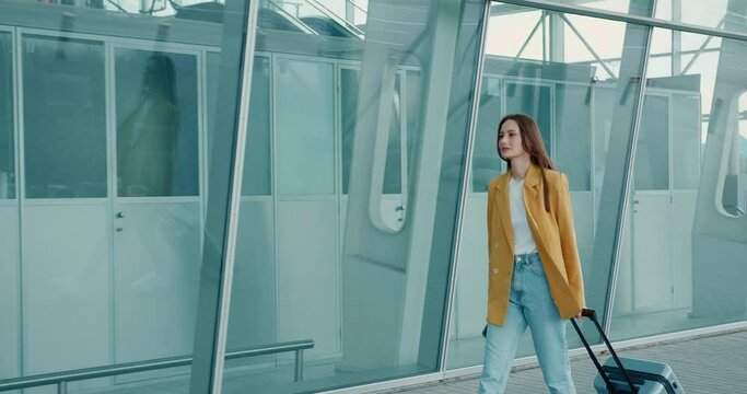 Adorable young woman in yellow jacket and blue jeans walking with suitcase and smartphone near modern airport. Concept of business trip. 