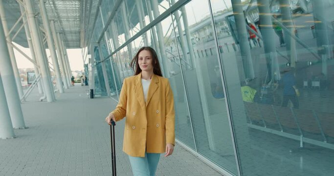 Beautiful young woman with brown hair walking near modern airport with gray suitcase. Business lady in yellow jacket and blue jeans having business trip. 