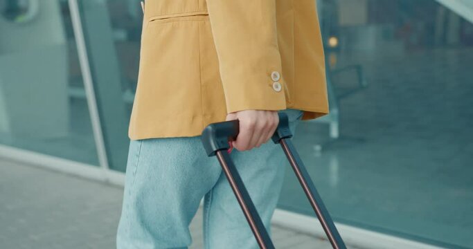 Close up of young woman wearing trendy yellow jacket and blue jeans walking near airport with suitcase. Concept of people and travelling. 