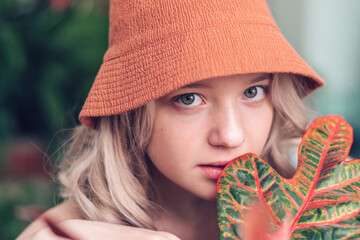 Portrait of young blonde girl in panama hat among the orange leaves of the codieum.Natural woman beauty.Closeness to the nature.Eco,urban jungle,biophilia concept.Skin care and wellness concept.