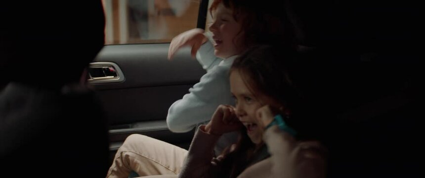 Cute little siblings brother and sister singing and going crazy on a back seat of a modern SUV while riding through neighborhood. Shot with 2x anamorphic lens