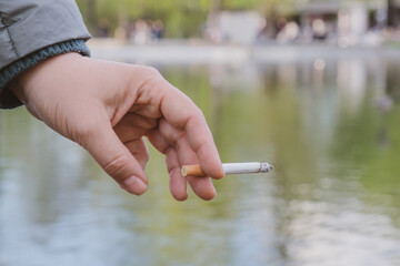 Female hand holds a burning cigarette against the backdrop of pond in a city park. Tendency to smoke. Dealing with emotional stress with smoking. Space for text.