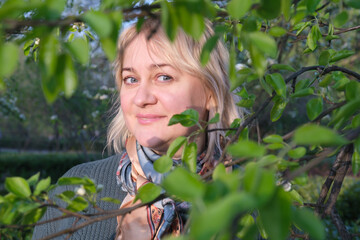 Portrait of a middle-aged woman with blond hair through tree branches with leaves. Flirty look and a light smile. A walk in spring park, romantic date, woman in love.