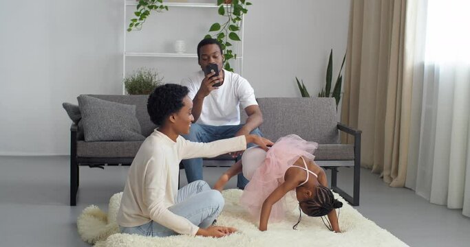 Afro american family spend time at home, african father sits on couch looking at mobile phone, black ethnic mother plays with girl daughter in pink dress doing acrobatic poses on floor on soft carpet
