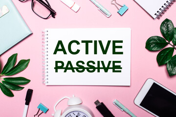 Fototapeta na wymiar The word ACTIVE is written in a white notebook on a pink background surrounded by business accessories and green leaves.