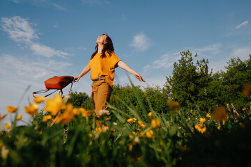 A young dark-haired woman in a yellow dress, with open arms, walks across the field looking at the blue, sunny sky. Wildflowers, summer time.