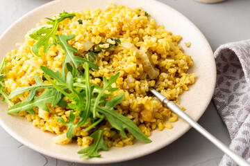 Delicious bulgur porridge with fried onions and herbs in a plate with arugula, top view