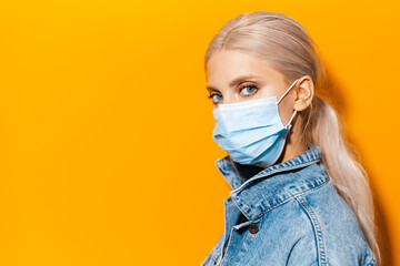 Side studio portrait of young blonde girl wearing medical face mask against coronavirus on background of orange color.