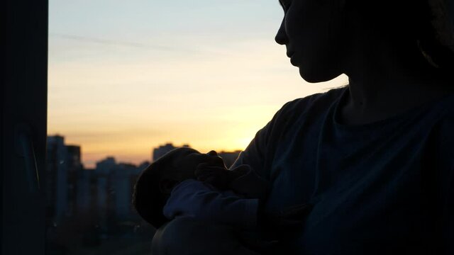 Mother hold newborn baby on hands, silhouetted shot against room window, sunset afterglow on sky. Moment of rest for tiny child, daughter lie quietly and look to mother