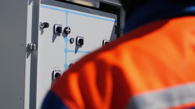 Worker hand in uniform opens door of cabinet with switchboard at distribution substation on sunny day close backside view