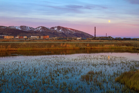 Evening arctic landscape. In the distance, an abandoned port town. View of the lake, tundra and mountains. There is a full moon in the setting sky. Beringovsky, Chukotka, Siberia, Far North of Russia.