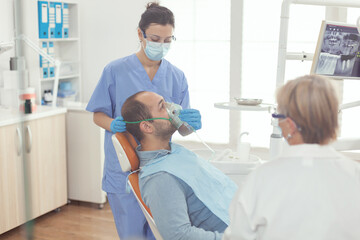 Obraz premium Medical nurse putting oxigen mask preparing sick man patient for stomatology surgery while sitting on dental chair in orthodontic hospital room. Dentist doctor examining toohache