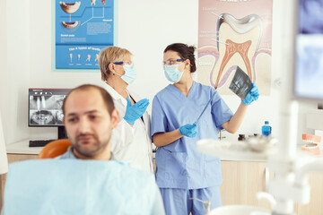 Stomatologist doctor and orthodontist assistant examining teeth radiography checking somatology problem while sick patient waiting on dental chair. Man preparing for surgery appointment