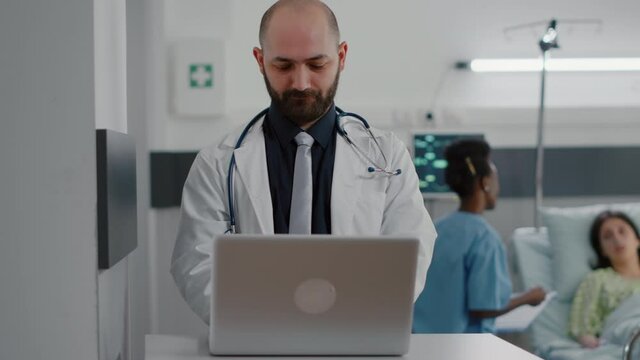 Front view of practitioner doctor typing illness expertise on computer while in background black nurse discussing healthcare treatment. Hospitalized patient having respiratory disorder