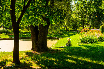 Woman in a hat sits on the shore of a pond in the park on a sunny morning in the shade of a tree