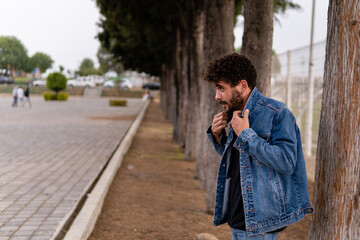 Handsome young man with curly hair and beard wearing denim posing and adjusting his jacket in a public park. Close up portrait with confident expression and copy space