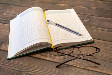 Opened diary, pen, and glasses on the brown wooden table closeup