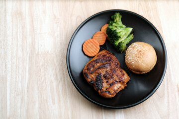 flat lay top view shot of homemade fried spicy chicken steak served with burger bread, sliced carrots, and pieces of broccolis in black dish on wooden table
