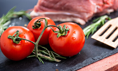 Fresh pork meat with ingredients for cooking on dark background. Tomato and rosemary