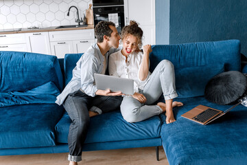 Happy family couple kissing and having fun on the sofa in a beautiful living room, smiling. Women is very happy for successful project. Joyful stylish husband and wife took a break from working.
