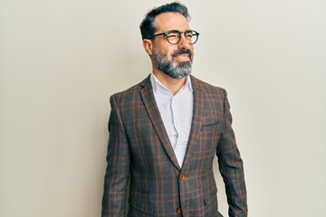 Middle age man with beard and grey hair wearing business jacket and glasses looking away to side with smile on face, natural expression. laughing confident.