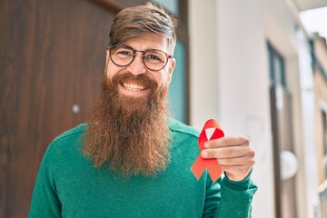 Young irish man with redhead beard smiling happy and holding red hiv ribbon at the city. - obrazy, fototapety, plakaty