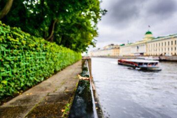 Rainy autumn day in the city, alley in the park near the river on which the ship is sailing. Close up view from the handrail on which lies the oak leaf on the embankment level
