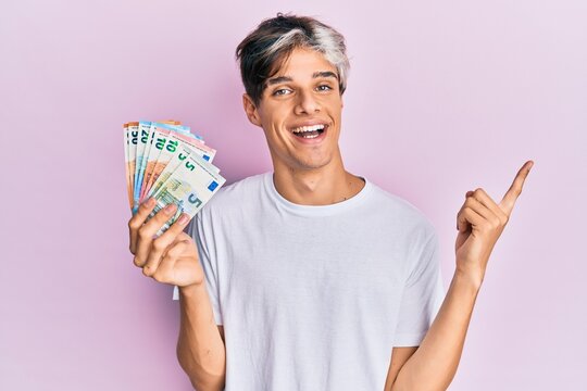 Young hispanic man holding euro banknotes smiling happy pointing with hand and finger to the side