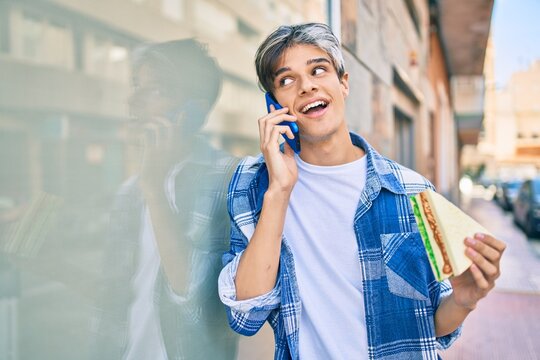 Young hispanic man smiling happy talking on the smartphone and eating sandwich at the city.