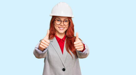 Young redhead woman wearing architect hardhat approving doing positive gesture with hand, thumbs up smiling and happy for success. winner gesture.