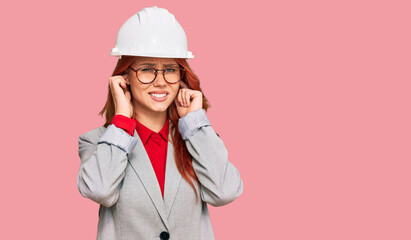 Young redhead woman wearing architect hardhat covering ears with fingers with annoyed expression for the noise of loud music. deaf concept.