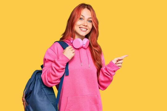 Young redhead woman holding student backpack and books smiling happy pointing with hand and finger to the side