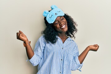 Beautiful african young woman wearing sleep mask and pajama very happy and excited doing winner...