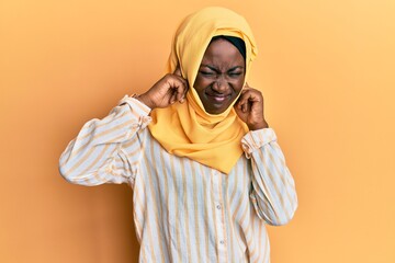 Beautiful african young woman wearing traditional islamic hijab scarf covering ears with fingers with annoyed expression for the noise of loud music. deaf concept.