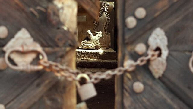 A locked lock hangs on a chain by old damaged gate with knockers, change focus at the damaged altar with sunlight on the fallen cross.