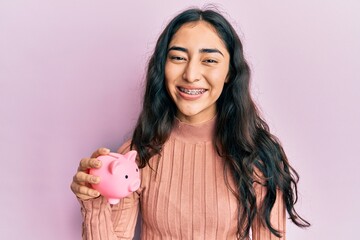 Hispanic teenager girl with dental braces holding piggy bank looking positive and happy standing and smiling with a confident smile showing teeth