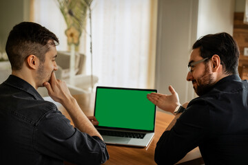 Zwei junge Männer, Anfang 20. sitzen im Wohnzimmer, schauen auf den Display eines Laptops. Einer zeigt mit den Fingern drauf,  der anderer  ist  nachdenklich. Green screen, grüner Bildschirm