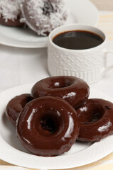 front, top view, close distance, of four freshly backed chocolate donuts covered with dark chocolate frosting on a round, white plate and a cup of black coffee