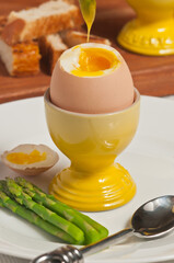 Front view, close distance of a soft boiled egg in a egg cup with the top cut off and an asparagus dipped into yolk on round white plate with four asparagus piers and an artisan spoon
