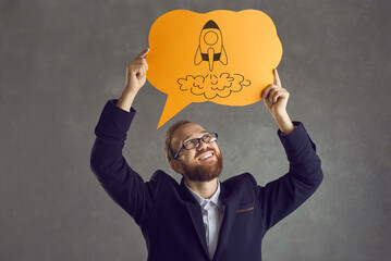 Smiling bearded man in suit holding paper thought bubble with spaceship doodle. Happy businessman in glasses looking at space rocket picture on speech balloon. Business, innovation, success concept - obrazy, fototapety, plakaty