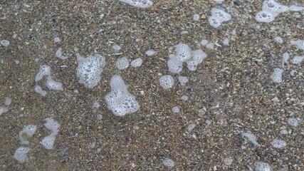 Unusual background of sand and small seashells filled with sea water with foam bubbles.