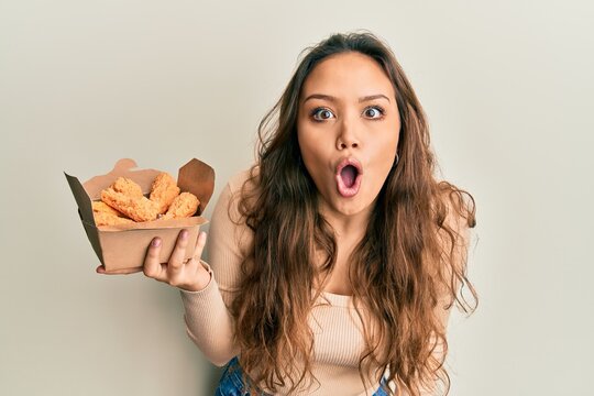 Young hispanic girl eating chicken wings scared and amazed with open mouth for surprise, disbelief face